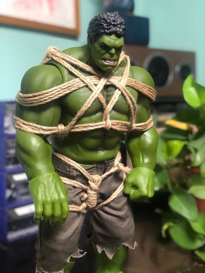 图片主体是一个绿巨人（Hulk）action figure，全身被粗麻绳以交叉缠绕的方式紧紧捆绑，绳索从胸膛延伸至腰部和手臂，形成密集的束缚效果。手办保持着标志性的绿色皮肤、隆起的肌肉线条和愤怒的面部表情——眉头紧锁、嘴巴大张露出牙齿，双拳紧握于身体两侧，仿佛正处于即将挣脱束缚的暴怒状态。下半身穿着标志性的灰色破洞短裤，裤腿边缘有撕裂痕迹。背景为室内居家环境，可见蓝色置物架、棕色音箱、绿色盆栽植物及墙上的挂画，整体场景具有生活气息。强大的绿巨人被普通麻绳捆绑的反差画面产生了幽默效果。