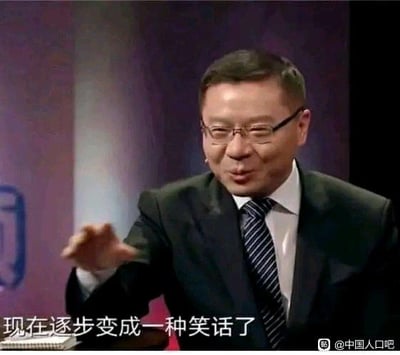 一位戴着眼镜、穿着黑色西装和条纹领带的男子，正坐在类似访谈节目的场景中，右手伸出做手势，表情略带无奈。图片底部配有文字：'现在逐步变成一种笑话了'，整体给人一种对某事表达不满或嘲讽的感觉。