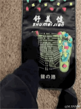 这是一段实拍动态图，画面中一只穿着深色袜子的脚踩在印有「舒美健 SHUMEIJIAN」字样的足部穴位按摩垫上，垫子上印有足部反射区示意图和「健の路」字样，脚在垫子上的动作显得有些滑稽，呈现出日常尝试足部按摩的生活化场景。