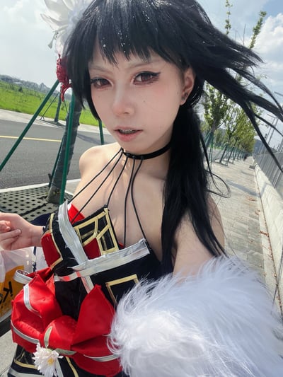 这是一张竖版的户外自拍照，画面主体是一名coser正在自拍，其cos的是音游《maimai》中的角色黑姬：佩戴黑色齐刘海长直发假发，假发上装饰有白色花朵，佩戴红色美瞳，妆容贴合角色设定，脖子上戴着黑色系带项圈，身上的cos服以黑、红、金三色为主，上身有黑色细挂带设计，腰部搭配大红色蝴蝶结装饰，手臂处配有蓬松的白色毛绒装饰。拍摄背景是晴朗白天的户外公共道路区域，身后可见人行道、行道树、沥青马路和远处的绿色草坪，整体光线明亮。