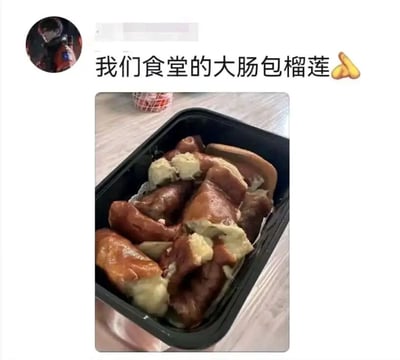 图片展示了一则社交媒体分享内容，上方有用户头像（已模糊处理）和文字“我们食堂的大肠包榴莲🍟”，下方是一张实拍图：黑色餐盒中装着几块棕色的大肠，内部包裹着黄色的榴莲果肉，整体呈现出一种奇特的食物组合，疑似食堂推出的创意（或黑暗）料理。