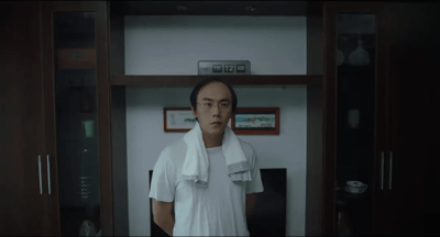 图片中是一名身着白色T恤、颈挂毛巾的男子，正站立在带有玻璃门的木质柜子前。柜子上层摆放着标有“闽D·12”字样的车牌及相框，背景中可见中式风格的装饰画与瓷器摆件，整体场景呈现出生活化的居家环境。该画面展现了“龙王赘婿”系列广告中“卑微赘婿”阶段的经典形象，人物神态平静中透着隐忍，是后续“身份揭露、打脸逆袭”剧情的铺垫。
