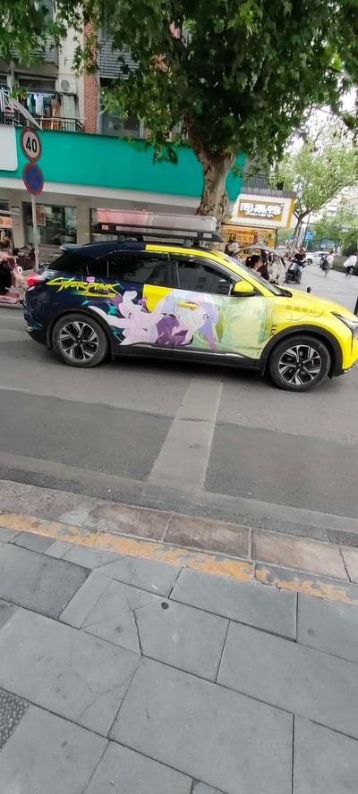 这是在国内城市街边拍摄的一辆个性化改装痛车，车辆为黄蓝撞色的新能源SUV，车身侧面涂装了动画《赛博朋克：边缘行者》中的人气角色露西，同时印有英文“CYBERPUNK”字样，车顶加装了行李架。车辆停在设有40km/h限速标志的街道旁，背景可见行道树、带有“周黑鸭”招牌的商铺，将二次元ACG文化与日常出行车辆结合，充满个性潮流感。