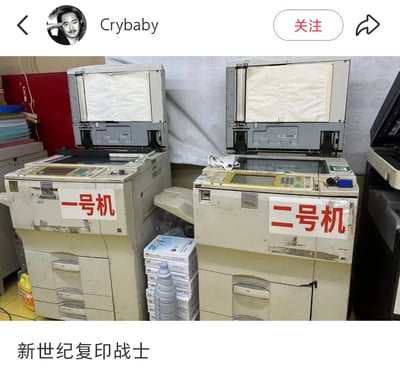 图片中展示了两台略显陈旧的理光（RICOH）复印机，左侧机器贴有红色“一号机”标签，右侧为“二号机”标签。两台设备机身有明显使用痕迹，顶部纸盒均处于打开状态，周围堆放着打印纸、清洁液瓶等耗材，整体呈现出办公室常见的设备使用场景。图片底部配有文字“新世纪复印战士”，巧妙化用了经典动画《新世纪福音战士》的名称，将老旧复印机调侃为“战士”，形成幽默反差效果。