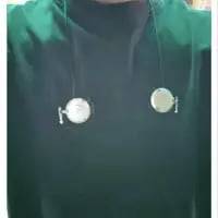 这是一张利用视觉错位拍摄的创意照片，图中真人穿着深绿色上衣，用细绳将两个磁带悬挂在脖子两侧的位置，通过精准的摆放制造出视觉错觉，让原本普通的磁带看起来像是从肩膀处延伸出来的身体部件，利用日常物品打造出趣味十足的创意视觉效果。