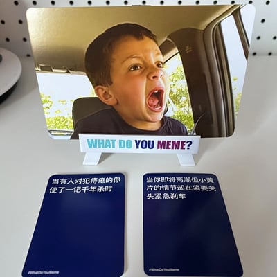 图片展示了What Do You Meme?游戏的中文版本，中央是一张印有惊讶男孩（网络迷因人物）的卡牌，男孩坐在汽车后座，表情极度震惊，嘴巴大张，眼睛圆睁。卡牌下方有两个蓝色的文字卡牌，分别写着："当有人对犯痔疮的你使了一记千年杀时"和"当你即将高潮但小黃片的情节却在紧要关头紧急刹车"。整个场景展示了这款派对游戏的玩法，即通过搭配图片和文字卡牌创造幽默效果。