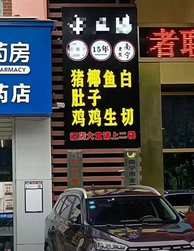 这是一张街头实拍的静态图片，画面主体是南宁一家有着15年经营标识的餐饮店铺竖版黑色招牌，招牌上的菜品文字因排版问题出现了歧义断句：原本应为“猪肚鸡、椰子鸡、鱼生、白切鸡”的菜品，被排成了“猪 椰 鱼 白；肚子 子 鸡 鸡；鸡 鸡 生 切”的形式，容易让人产生误解，形成搞笑效果。招牌下方标注“酒店大堂请上二楼”。画面左侧是一家蓝色的连锁药店，右侧是其他商业店铺，招牌下方地面有两根石柱，右侧石柱可见“南宁市金”字样，下方停着一辆深色SUV车辆。整个场景是日常的城市街头商业区域。