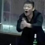 图片中一名穿着黑色上衣的男子，嘴巴大张呈呼喊状，双手合十做出用力鼓掌的动作，表情看起来极度激动或兴奋。背景隐约可见室内环境，桌子上放有一个绿色瓶子，整体场景可能是在聚会、饭局或类似的社交场合。该图常被用作反应表情包，表达强烈的赞同、兴奋、起哄或调侃等情绪。