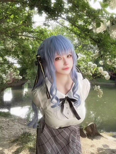 图中是一位打造了二次元风格造型的coser，戴着雾霾蓝色长卷发齐刘海假发，搭配黑色带金边的蝴蝶结发饰，身穿带有黑色领结的精致白色衬衫，下身搭配灰格纹下装。她背手站立在开满白色绣球花的树木下，背景是静谧的湖水，阳光透过枝叶缝隙洒落，整体画面清新柔和，充满治愈感，完美融合了二次元风格与自然实景。
无谐音梗、双关或暗示内容。