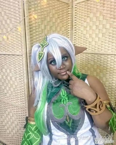 这是一张真人cosplay静态照片，一位黑人女性扮演了米哈游游戏《原神》中的草元素神明纳西妲（小吉祥草王）。她佩戴了还原角色的银绿渐变假发与精灵耳饰，身着带有草元素标志性三叶草、藤蔓纹样的绿白配色服饰，左手腕也戴着对应元素主题的臂饰。她用手指轻抵嘴唇，眼神直视镜头，神态略带俏皮。背景是编织纹理的墙面，上方挂着暖黄色的串灯，整体场景简洁，突出cos的角色形象。由于coser的外形与原作中纳西妲软萌娇小的浅色皮肤形象反差较大，这组cos照在网络上引发了关注与讨论。