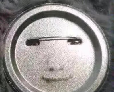 这是一张金属徽章背面的实拍图，原本是普通的徽章别针结构和污渍，却因为视觉错位形成了类似人脸的效果：横向的别针如同嘴巴，下方的深色污渍分别对应眼睛和鼻子，让原本平淡的物品背面看起来像一张表情呆滞的人脸，属于偶然出现的趣味视觉巧合。