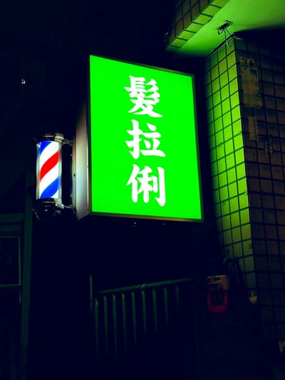 本图以夜间街拍形式，展现一家理发店门口的招牌。大大的绿色发光板上用繁体字写着“髮拉俐”，左侧还有理发店经典旋转柱。该词是网络流行谐音梗，模仿英文farali，给人土味和魔性质感，经常被网友拿来调侃理发店门牌，属于恶搞和创意网络文化。整个画面既复古又带点恶趣味，非常适合娱乐或玩梗场景。