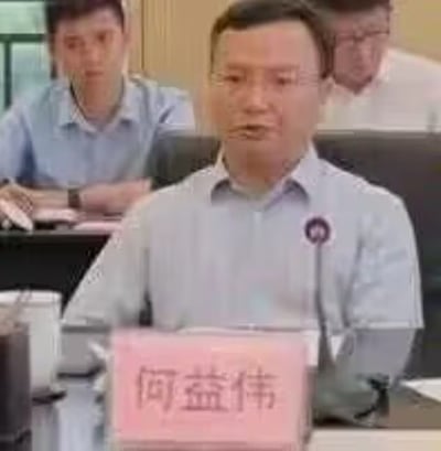 这是一张工作会议场景的实拍照片，画面核心人物是名为何益伟的男士，他穿着带有圆形标识的浅灰色衬衫，坐在会议桌前，神情专注，似乎正在发言或参与会议讨论。他面前的粉色姓名牌清晰标注着“何益伟”的名字，桌上还放置着水杯等会议常用物品。背景中能看到其他两位参会人员，整体场景是正式的室内办公会议环境，布置简洁规整，传递出严肃、专注的职场工作氛围。