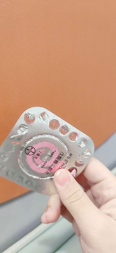 这是一张构图清晰的竖版原始拍摄图，画面中一只真实的人手拿着部分使用过的补佳乐（戊酸雌二醇片）铝塑药板，药板上可见剩余的粉色药片，中间区域标注了药品名称「戊酸雌二醇片」、品牌「补佳乐®」、规格「1毫克」以及生产厂商「BAYER 拜耳」的标识，还有产品批号、有效期至的相关标注区域，背景是纯色的暖橙色平面，清晰展示了这款妇科雌激素类药物的实物状态。