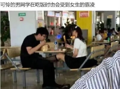 这是一张在学校食堂拍摄的搞笑错位照片，画面中一男一女各自坐在餐桌两侧吃饭，由于拍摄角度的巧合，女生的腿和男同学的腿在视觉上重叠，看起来像是女生把腿搭在男同学腿上，营造出“霸凌”的假象。图上方配文“可怜的男同学在吃饭时也会受到女生的霸凌”，利用视觉错位制造出反差感，带来滑稽搞笑的效果，没有双关、谐音梗等内容，纯靠错位画面和调侃文字形成笑点。