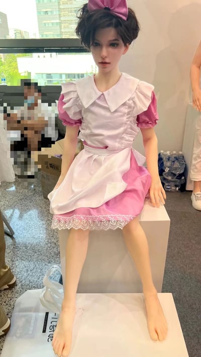 室内展览场景中，一个高度仿真的等身人偶坐在白色基座上。人偶有着精致的五官和深色短发，头上戴着一个大大的粉色蝴蝶结。上身穿着白色带有褶皱设计的短袖上衣，袖子为粉色泡泡袖，下身搭配粉色裙子，裙摆处有白色蕾丝花边装饰，整体呈现出可爱的女仆装风格。背景中可以看到其他展览物品、几位参观者以及窗外的城市建筑，地面上散落着一些展览相关物品。