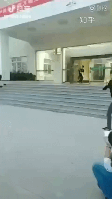 这是一段动态GIF，画面呈现了一栋带台阶的建筑入口场景：一名男子从建筑门内慌张地快速跑出，前景处有一人手持物品做出模仿射击的动作，模拟出类似追逐射击的趣味场景，整体氛围轻松滑稽，没有双关、谐音梗或暗示内容。