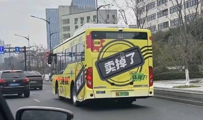 这是一张从车内视角拍摄的城市道路实拍图，画面中一辆黄色公交车十分醒目，车身被闲鱼平台的广告全覆盖，车尾和侧面都印有大号的‘卖掉了’字样，搭配‘拜拜了！’的趣味文字，还有闲鱼的品牌标识，以生活化的网感风格宣传闲置物品交易业务，显得接地气又有趣。