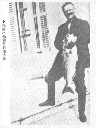 这是一张黑白纪实老照片，图中人物为托洛茨基，他身着深色外套与长靴，脸上带着笑容，双手紧紧抱着一条自己亲手钓获的大鱼，神态十分愉悦。图片左侧配有竖排繁体文字“托洛次基親手釣獲大魚”，明确标注了图中人物与事件内容。