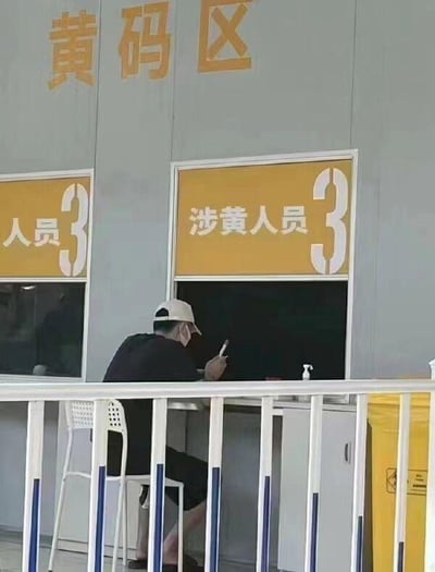 图片显示一个临时搭建的场所，上方有'黄码区'三个黄色大字。下方有两个窗口，左侧窗口标有'人员3'，右侧窗口标有'涉黄人员3'。一名戴着白色帽子和口罩的男子正坐在右侧窗口前使用手机。

图片存在明显的文字歧义问题：'涉黄人员'在中文语境中通常指涉及色情活动的人员，与防疫场景中的'黄码人员'概念混淆，可能是由于标识设计不当造成的误解。