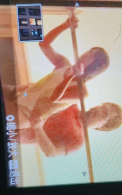 这是一张家用监控设备界面的翻拍截图，画面中呈现两个孩童共同抓握着一根竖直的杆子，看起来正在玩耍。界面左上角设有多画面监控的分屏预览小窗口，左下角带有“缩小/放大”“退出”的操作按钮以及设置图标，整体画面存在一定的模糊感，是典型的监控设备操作界面。