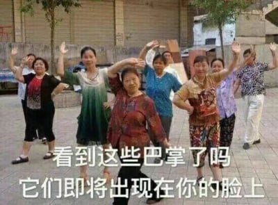 这是一张具有搞笑戏谑风格的表情包，画面主体是一群在户外砖地空地上跳广场舞的中老年女性，她们穿着日常休闲服饰，大多面带笑容，抬手摆出舞蹈动作，姿态轻松活泼。图片下方配有两行文字“看到这些巴掌了吗 它们即将出现在你的脸上”，将大妈们跳舞时抬手的动作夸张调侃成要扇巴掌的威胁姿态，通过日常场景和“放狠话”文字的反差感制造出强烈的搞笑效果，适合在和朋友开玩笑、吐槽不满、幽默放狠话的社交场景中使用。