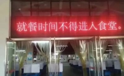 这是一张拍摄食堂入口的实拍图，食堂门口的红色电子显示屏上标注着「就餐时间不得进入食堂」的规定标语。这一规定完全违背食堂的使用逻辑，就餐时间本是人们进入食堂用餐的时段，这种自相矛盾的内容制造出荒诞搞笑的迷惑效果。