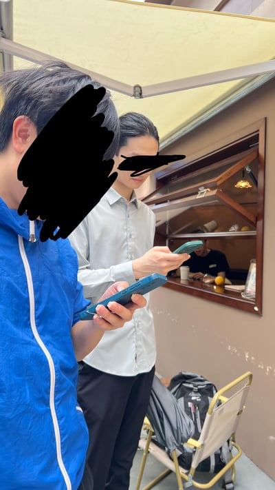 这是一张竖拍的真实日常场景照片，画面中两位男士站在带有遮阳棚的街边小店窗口前，均低头专注于手中的手机，两人的面部被做了马赛克处理以保护隐私；窗口内侧坐着一位男士，看起来像是店铺的工作人员，整体氛围平淡日常，展现了现代人随时随地使用手机的状态。