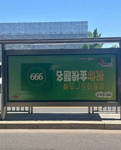 这是一张户外公交站台的实拍照片，画面主体是一块被反向悬挂的999感冒灵户外广告牌，正常视角看到的所有文字都是倒置状态，将内容正过来后可识别为“故意挂反广告牌 祝你金榜题名”，同时配有倒置的999品牌标识、感冒灵产品图案、“暖暖的很贴心” slogan以及OTC广告标识。该创意利用“反挂”的寓意，暗含高考考生不会挂科的美好祝愿，搭配金榜题名的祝福文字，是贴合高考节点的趣味营销内容。