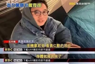这是一张中国台湾东森新闻的视频截图，画面中是一位在高雄户外彻夜排队的民众，他裹着厚外套，身处帐篷旁，看起来状态疲惫。图中的新闻字幕玩了一个调侃的梗：民众为了购买售价高达九万元的高端显卡彻夜排队，字幕戏称“手机拿起来 看黄仁勋的照片 身体就暖起来了”。黄仁勋是NVIDIA的创始人兼CEO，该品牌是高端显卡的主流厂商，这个梗用夸张的方式调侃了显卡爱好者对NVIDIA的狂热信仰，以及彻夜排队的辛苦，靠精神力量“取暖”，充满搞笑和戏谑的意味。