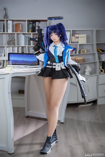 这是一张真人cosplay摄影作品，coser还原了手游《蔚蓝档案》中的角色早濑优香：深蓝色双马尾发型搭配标志性的悬浮光环，身穿蓝白黑配色的制服套装，双手各持一把带有蓝色装饰的仿真枪械，在摆放着电脑、书籍的办公室场景中摆出干练的战斗姿态，完美还原游戏中角色的冷峻干练气质。