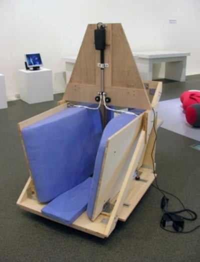 画廊场景中展示着一件实验性装置作品：主体为木制框架结构，顶部呈三角形，中央垂直安装有金属杆及黑色部件，底部设有蓝色软垫座椅，侧面可见连接的电线。装置放置于灰色地面，背景有多个白色展台，其中一个展台上摆放着小型显示器（屏幕显示模糊内容），右侧地面上有红色与灰色的物体。整体呈现出艺术与科技结合的实验性风格，可能是交互式装置或概念设计原型。