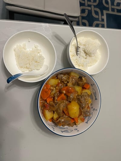 这是一张俯拍视角的家庭餐食照片，呈现出一顿温馨的家常饭：画面里有两碗白米饭，左侧碗中的米饭已被食用一部分，搭配一把带蓝纹的白瓷勺；右侧碗里的米饭完整，插着一把金属勺子。中间的碗中装着炖煮入味的土豆胡萝卜炖肉，肉块、粉糯的土豆块、胡萝卜块裹着浓郁酱汁，看起来十分下饭，整体场景充满日常家庭用餐的温暖感。