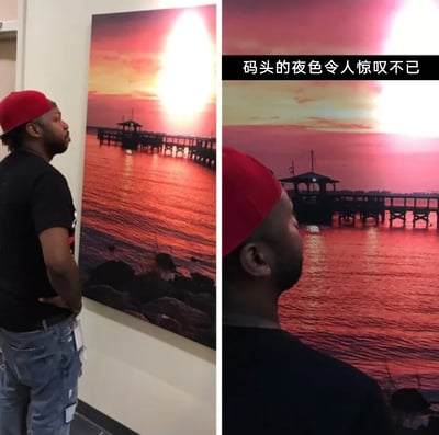 这是一组左右拼接的对比图，左侧图中，一位戴着红色帽子、身穿黑色上衣和破洞牛仔裤的黑人男子背对着镜头，站在室内环境中，正注视着墙上悬挂的一幅码头日落主题装饰画，画中是橙红色调的海面、延伸的码头栈桥与耀眼的落日；右侧图通过调整拍摄视角，将男子的头部置于画面下方，与装饰画的内容巧妙结合，营造出男子仿佛真实站在画中的码头前，正在观赏眼前日落海景的趣味视觉错觉效果，画面右上角配有文字“码头的夜色令人惊叹不已”，进一步强化了这个充满创意的视觉错位梗，整体效果诙谐有趣，利用构图制造出逼真的场景混淆感。