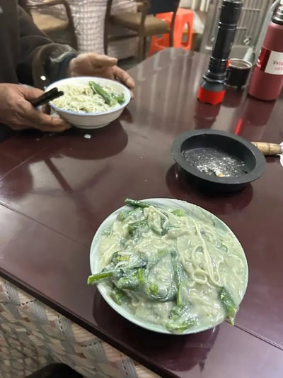 图片展示了两碗面食，但前景中的那碗面异常粘稠，看起来像是淀粉浆糊混合了蔬菜和面条，整体色泽发白，非常诡异，令人食欲全无。背景中一只手正拿着另一碗普通的面条作对比，桌上还有喝水杯、手电筒和烟灰缸，环境显得接地气、朴素。这种极度稠糊的面条容易让人联想到失败的烹饪或“黑暗料理”，可能被网友戏称为“黑暗料理现场”或“厨房灾难”，带有浓厚的搞笑与无语气氛。没有发现直接的双关、谐音梗。