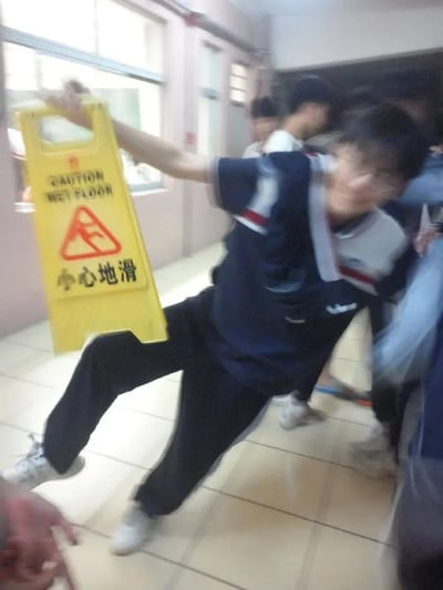一名穿着蓝白相间校服的学生，在学校走廊的瓷砖地面上，右手高举着黄色的“CAUTION WET FLOOR 小心地滑”警示牌，身体却向后倾斜，左腿向前伸出，呈现出即将滑倒的动态姿势。背景中可见其他模糊的学生身影和走廊的门窗，整体画面因运动产生轻微模糊，突出了“拿着防滑警示牌却自己险些滑倒”的滑稽反差感。