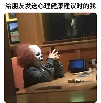 图片上方配有文字“给朋友发送心理健康建议时的我”，下方是一名戴着白色小丑面具、顶着红色卷发的人，坐在木质桌子前，双手合十置于脸前，姿态像是在认真地给出建议。背景为室内环境，有木质墙面和一台蓝色屏幕的设备。图片通过小丑面具的滑稽形象与“提供心理健康建议”的严肃行为形成反差，营造出幽默讽刺的效果，调侃了人们在给朋友提建议时可能显得一本正经却又略带滑稽的状态。