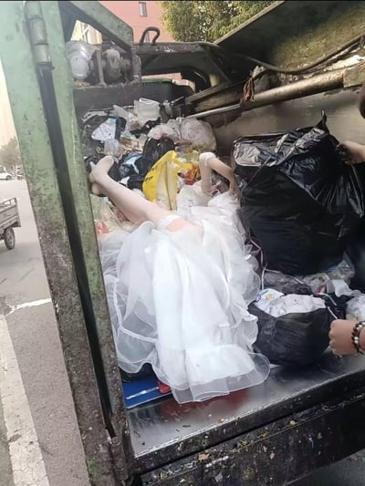 在城市街道旁的垃圾清运车车厢内，堆满了各色垃圾袋与杂物，中间躺着一个身着白色婚纱的人偶。人偶的腿部和手臂摆放角度极具迷惑性，乍一看会让人产生“真人被丢弃在垃圾中”的错觉，属于典型的视觉错位场景。画面右侧还有人正在整理黑色垃圾袋，背景能看到街道和车辆，整体场景充满了反差感，无谐音梗、双关等隐含内容，仅靠视觉错觉制造趣味效果。