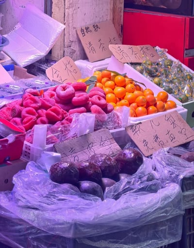 图片展示了一个充满生活气息的街头水果摊，摊位上陈列着多种水果：粉红色的果实（疑似柿子）、橙黄色的金桔、深紫色的水果（可能是李子或软儿梨），以及部分用透明包装纸包裹的绿色果实。摊位上方和水果旁摆放着多张手写 cardboard 招牌，内容包括“金桔”、“自然熟 清茶桔”、“清茶桔”、“软儿梨 免费真空”等字样，字迹质朴。摊位背景可见红色柜子、塑料包装盒和白色泡沫板，整体环境带有市井的质朴感。灯光呈现出淡淡的紫色调，为水果增添了鲜艳的色彩对比，展现了典型的街头市场风貌。