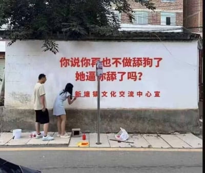 这是一张实拍图，画面里一面白墙上写着新塘镇文化交流中心宣传的扎心标语：“你说你再也不做舔狗了
她逼你舔了吗？”，墙前站着一男一女，女生正弯腰在墙面前操作，旁边放着墙绘用的水桶、颜料等工具。标语用年轻人熟悉的网络热词，直接戳破了部分人以“被迫”为借口自我感动式的“舔狗”行为，官方宣传用如此接地气的玩梗方式，反差感拉满，让人忍俊不禁。