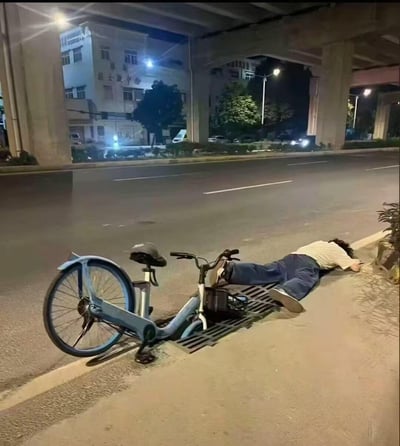 深夜的城市高架下方的道路边，一名男子面部朝下趴在人行道边缘，身旁倒着一辆共享单车，看起来像是因疲惫或醉酒失去力气倒下，画面展现了都市普通人的疲惫瞬间，满是生活的无奈感。