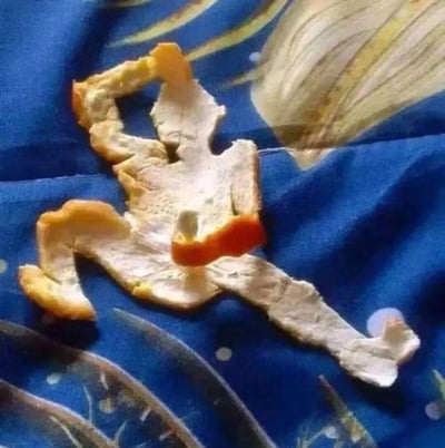 这张图里，一块吃剩的橘子皮被巧妙摆成了类似人类做劈叉动作的造型：一条腿伸直打开，另一条腿弯曲，一只手臂抬起，姿态生动逼真，仿佛正在做一个充满张力的动作，放置在带有装饰图案的蓝色布料上，充满了生活里的小创意，让人忍俊不禁。