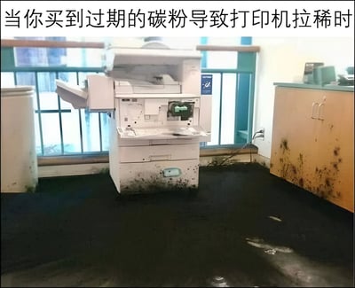 图片展示了一台白色办公打印机因使用过期碳粉而发生严重碳粉泄漏的搞笑场景。打印机机身及周围地面、柜面布满黑色碳粉污渍，如同“拉稀”般狼狈不堪。顶部文字“当你买到过期的碳粉导致打印机拉稀时”以拟人化比喻夸张呈现故障后果，营造出职场生活中的荒诞幽默。场景疑似办公室或公共打印区域，背景可见窗户护栏与浅色柜体，整体画面脏乱与文字的诙谐描述形成强烈反差。