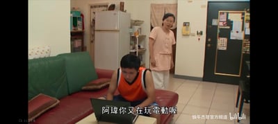 图中展示了一个家庭场景，一名穿着橙色背心的年轻男子（阿璋）正坐在红色沙发上专注地使用笔记本电脑。他的面前放着电脑，神情投入。在他身后，一位穿着粉色上衣和白色裤子的中年女性正站着，表情略带无奈地看着他。房间内有绿色的沙发、红色的靠垫、白色的冰箱以及各种家居用品，营造出生活化的氛围。图中下方有字幕“阿璋你又在玩電動喔”，暗示中年女性对年轻人沉迷游戏的不满。