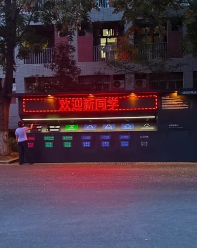 夜晚的校园场景中，一座垃圾分类站格外醒目。顶部红色LED显示屏滚动显示“欢迎新同学”字样，下方整齐排列着多个不同颜色的分类垃圾桶，分别对应可回收物、厨余垃圾等类别。一名身穿白色上衣的人正站在垃圾桶旁，似乎在投放垃圾或查看分类说明。背景是一栋多层教学楼，部分窗户透出灯光，周围有树木环绕，营造出温馨的校园氛围。