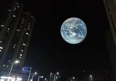 这张图片展示了一个超现实的城市夜景场景。画面中，两栋高耸的摩天大楼矗立在两侧，中间的夜空中悬浮着一个巨大的地球。地球的细节清晰可见，蓝色的海洋和白色的云层形成鲜明对比，仿佛近在咫尺。地面上可以看到街道的灯光和路灯，与上方巨大的地球形成强烈的视觉反差，营造出一种科幻感和震撼效果。