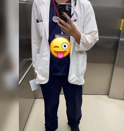 图片中是一位穿着白色医生大褂和深蓝色手术服的人，脖子上挂着听诊器，胸前有一个透明证件牌。此人正站在电梯里用手机自拍，手机壳上有黑色底和白色斑点图案。其面部被一个黄色搞怪emoji覆盖，该emoji左眼眯起 wink，右眼睁大，伸出粉色舌头，表情夸张滑稽。电梯内部为金属材质，可见银色的电梯壁和扶手，地面是浅色瓷砖。