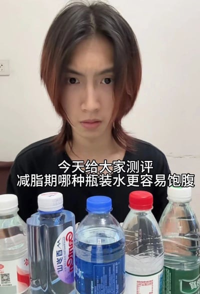 图片中一位短发人士面前摆放着五瓶不同品牌的瓶装水，包括百岁山、农夫山泉等常见品牌。人物表情严肃，正对着镜头，图片中间有文字：'今天给大家测评减脂期哪种瓶装水更容易饱腹'。这是一张带有明显幽默讽刺意味的图片，因为水本身没有热量，不存在'饱腹'效果，通过一本正经测评无意义内容制造笑点。