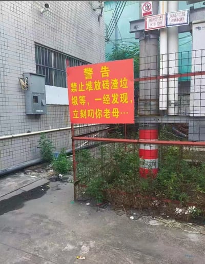 这是一张实拍的城市闲置角落照片，画面中心立着一块红底黄字的警告告示牌，告示牌内容为“警告 禁止堆放砖渣垃圾等，一经发现，立刻叼你老母…”，使用粗俗脏话作为违规警告内容。告示牌固定在红框铁丝网围栏上，背景是铺着灰色瓷砖的建筑墙面，墙面上设有玻璃窗和灰色电箱，围栏内长满杂草，地面散落少量垃圾，整体是城区无人管理角落的场景。
