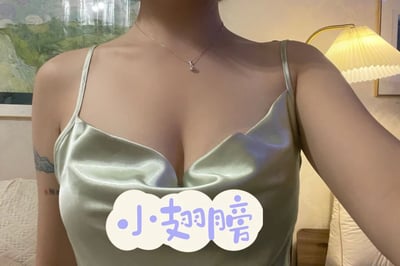 一位女性的自拍特写，展示了她穿着一件浅绿色缎面吊带衫的上半身。衣服采用深V领设计，凸显出优雅的锁骨线条。她佩戴着一条精致的细项链，吊坠小巧精致。左臂上有一个小纹身，增添了个性元素。背景中可以看到一盏带有褶皱灯罩的台灯和墙上的装饰画，营造出温馨的室内氛围。图片底部中央有一个云朵形状的文字贴纸，上面写着'小翅膀'三个蓝色汉字，为整体画面增添了可爱感。